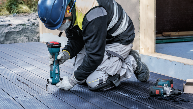 A person wearing safety equipment uses a cordless tool to cut decking boards.