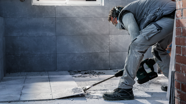 Person wearing safety equipment removes floor tiles using a power tool.