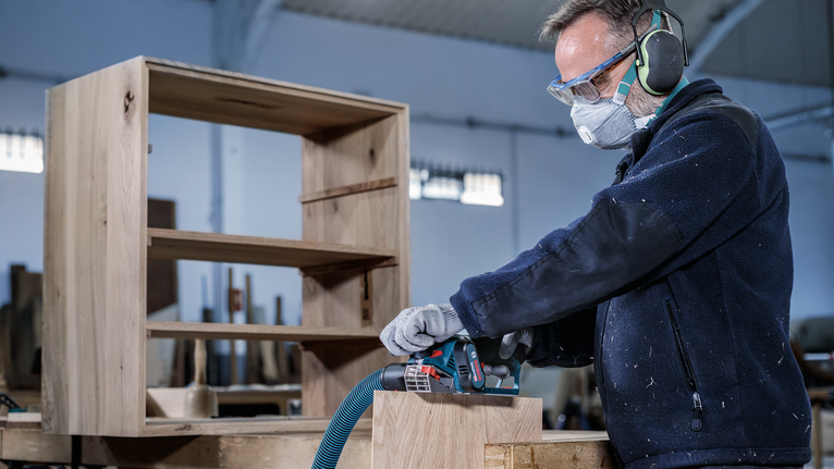 A person wearing safety equipment sands wooden furniture with a power tool.