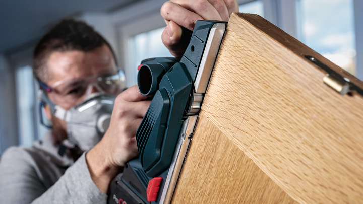 Person wearing safety equipment planes the edge of a wooden door with a power tool.