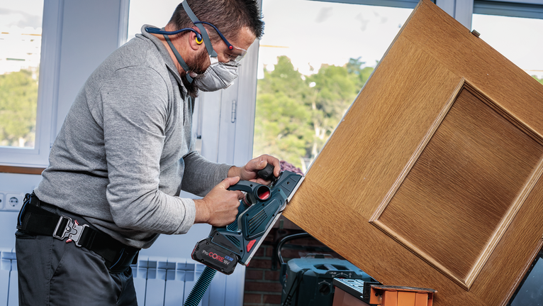 A worker wearing safety equipment sands a wooden door with a cordless power tool.