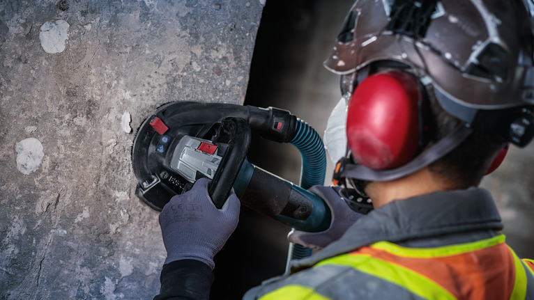 Person wearing safety equipment grinds concrete wall with a power tool.