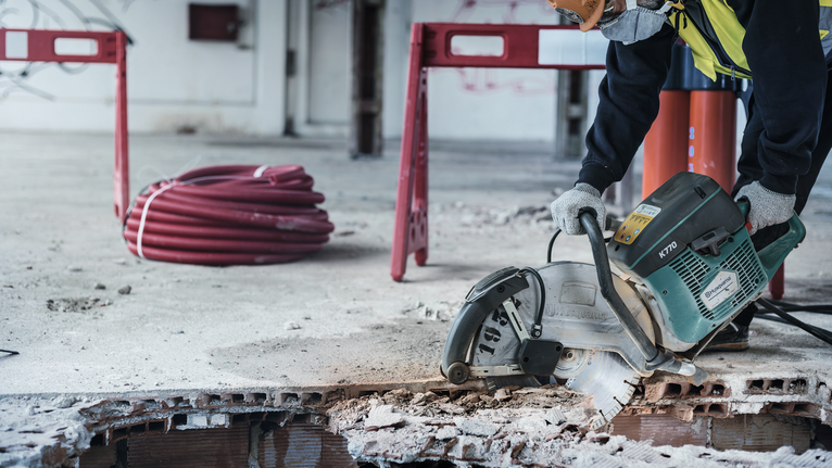 Worker wearing safety equipment uses a power cutter to saw through concrete flooring.