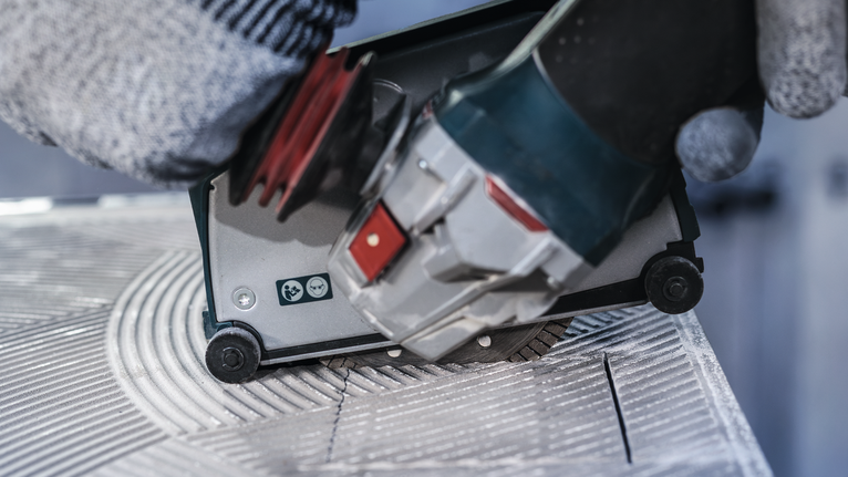 Person wearing safety equipment guides a power tool to cut grooves in a tile.