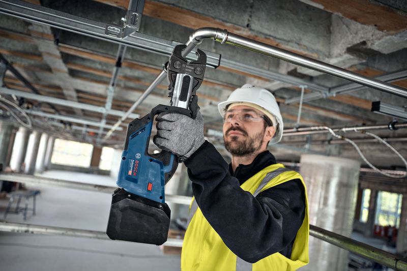 A person wearing safety equipment uses a cordless press tool on a metal pipe.