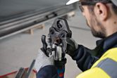 A person wearing safety equipment operates a cordless press tool on a metal fitting.