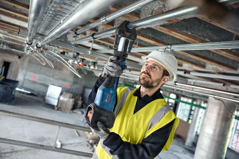 A person wearing safety equipment presses a pipe joint using a cordless press tool overhead.