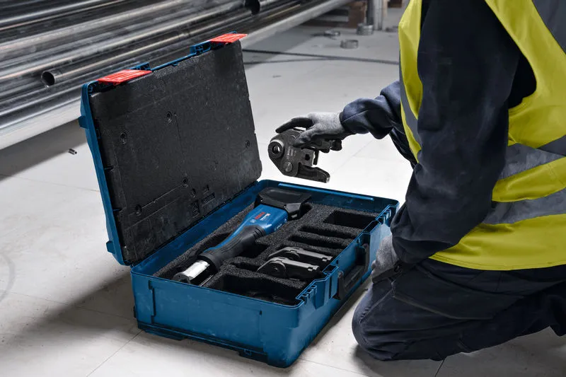 A person wearing safety equipment examines a cordless press tool in a blue carrying case.