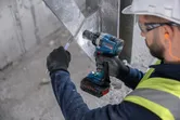 A person wearing safety equipment uses a cordless impact driver on a metal beam.
