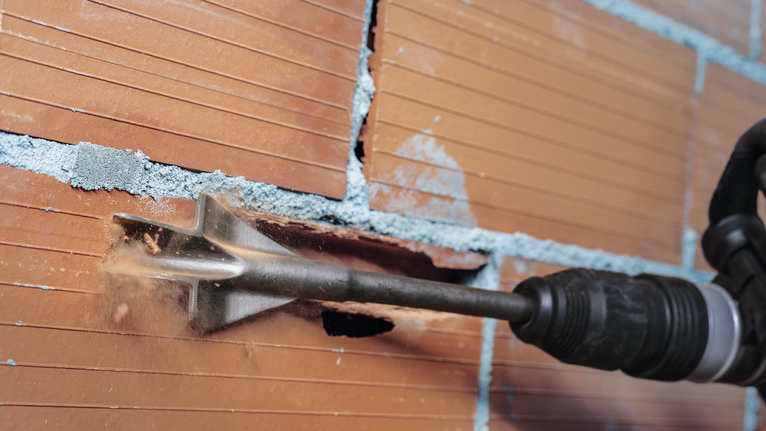 Chisel tool removing mortar between bricks in a masonry wall.