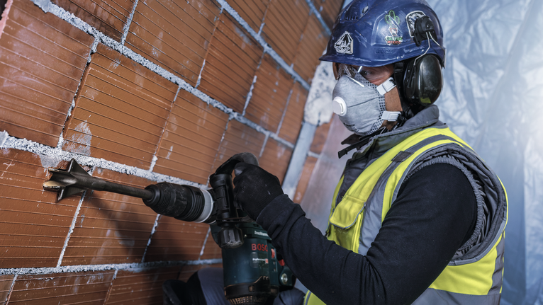 A person wearing safety equipment drills into a brick wall with a power tool.