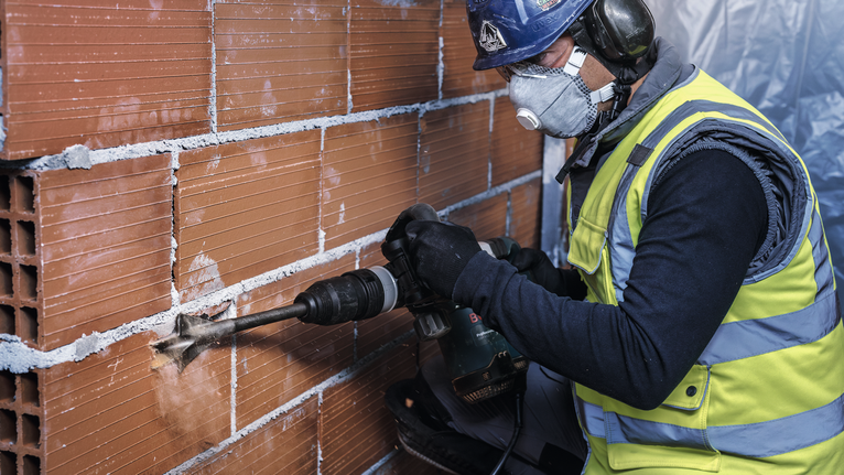 Person wearing safety equipment drills into a brick wall.