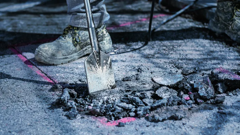 Person wearing safety equipment breaks asphalt with a demolition tool.