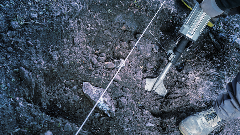 Person wearing safety equipment breaks soil in a pit using a demolition tool.