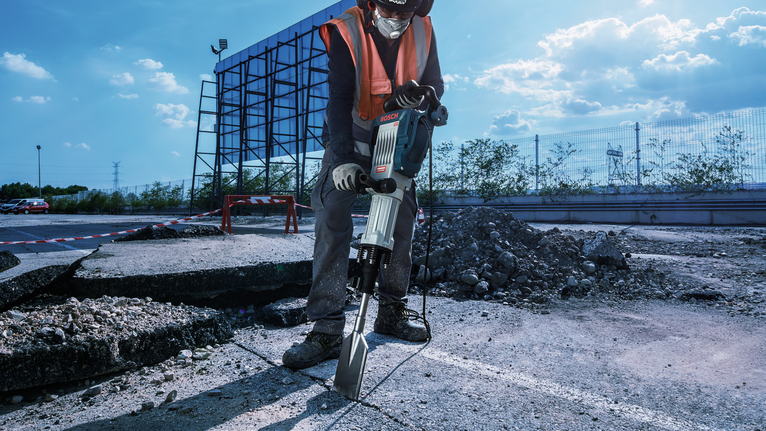 A person wearing safety equipment operates a demolition hammer to break asphalt pavement.