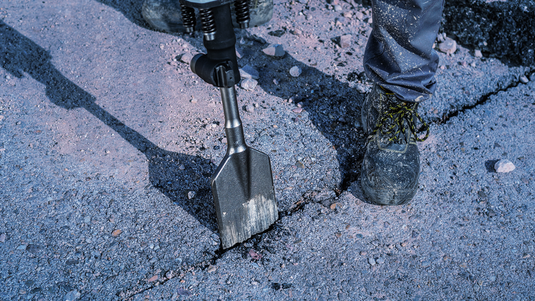 Worker wearing safety equipment uses a demolition hammer to break concrete pavement.