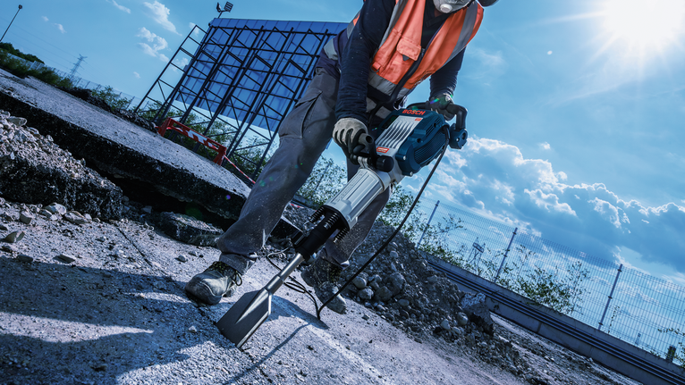 A person wearing safety equipment uses a jackhammer to break concrete on a construction site.