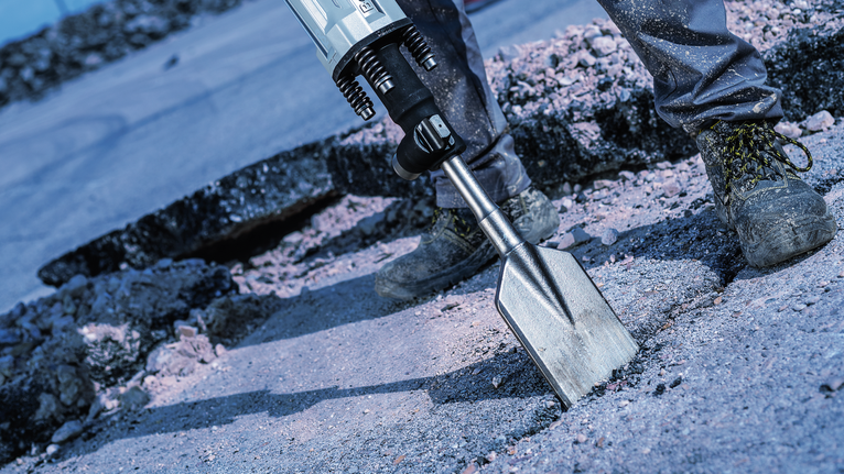 Person wearing safety equipment uses a demolition hammer to break concrete.