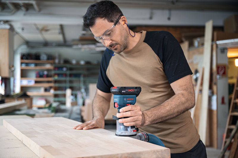 A person wearing safety equipment routes the edge of a wooden board with a cordless palm router.