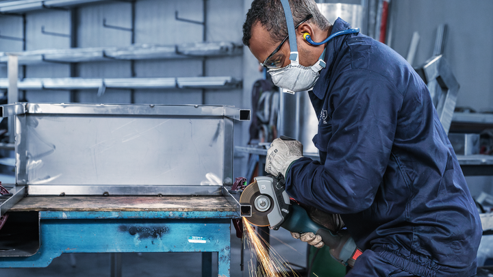 A person wearing safety equipment grinds metal, producing sparks in a workshop.