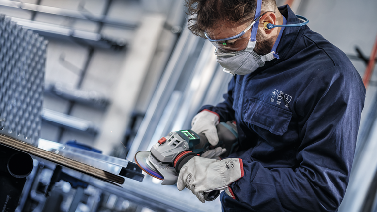 A person wearing safety equipment grinds metal with an angle grinder.