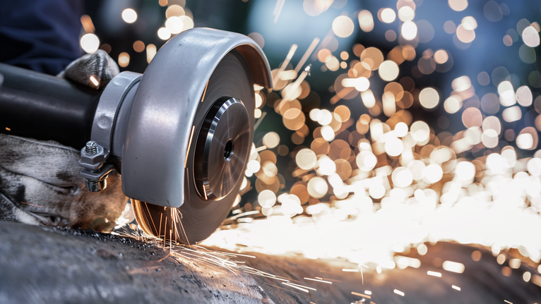 Person wearing safety equipment uses an angle grinder to cut metal, creating sparks.