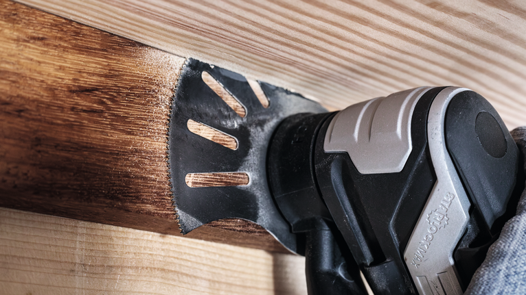 Person wearing safety equipment uses a multi-tool to sand wooden beams.