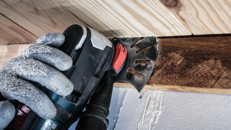 Person wearing safety equipment cuts wood with a multi-tool near a ceiling.