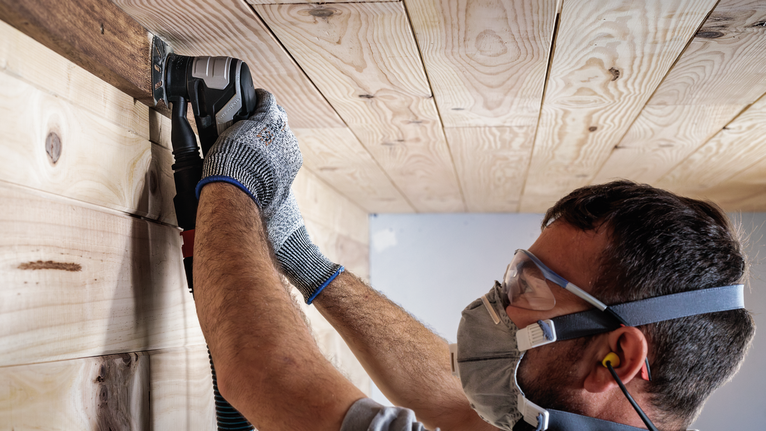 Person wearing safety equipment sands a wooden ceiling with a power tool.