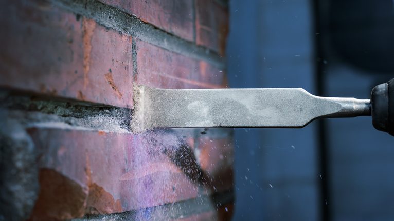 Chisel removes mortar from brick wall, dust scattering.