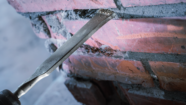 Chisel bit removing mortar from between red bricks.
