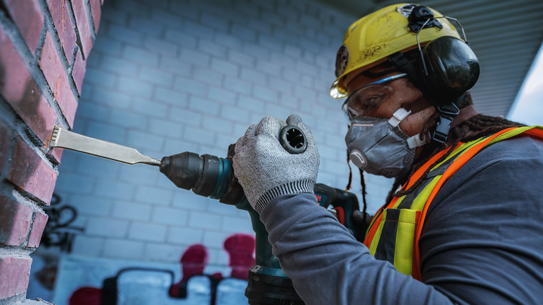 Person wearing safety equipment uses a power chisel to remove mortar from a brick wall.