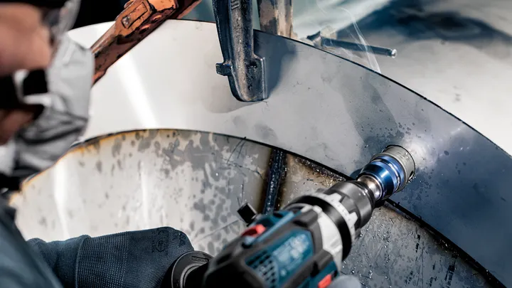 Person wearing safety equipment drills a hole into a curved metal surface.