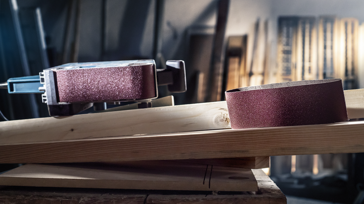 Belt sander and sanding belt placed on a wooden plank in a workshop.