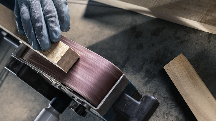 Person wearing safety equipment sands a wooden block on a belt sander.