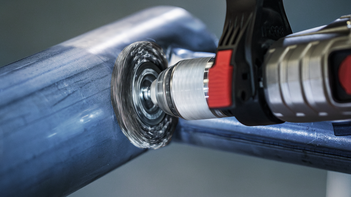 Power drill with a wire brush attachment sanding a metal pipe.