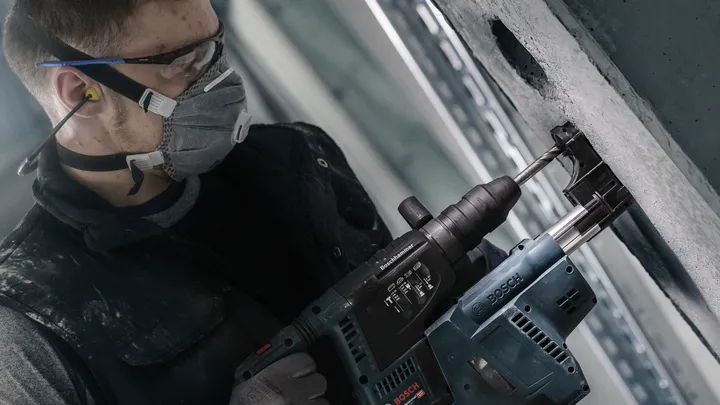 A person wearing safety equipment drills into a concrete wall with a power tool.