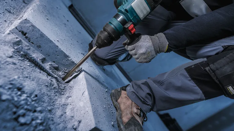 A person wearing safety equipment drills into concrete using a rotary hammer.