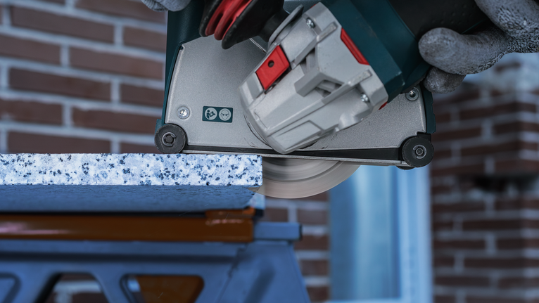 Person wearing safety equipment cuts a stone slab with a power saw.