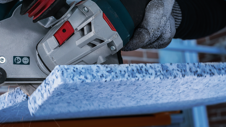A person wearing safety equipment cuts a blue speckled stone slab with a power tool.