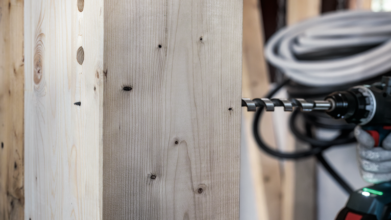 Person wearing safety equipment drills a thick wooden board with a power tool.
