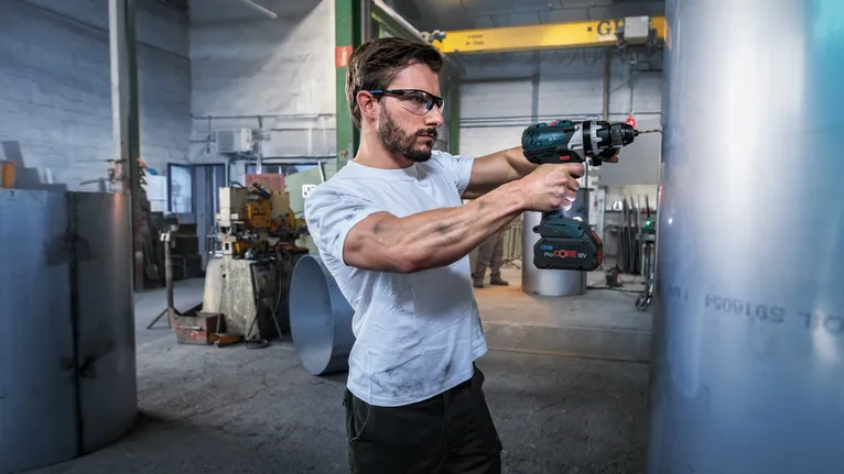 Person wearing safety equipment drills into a large metal cylinder in an industrial workshop.
