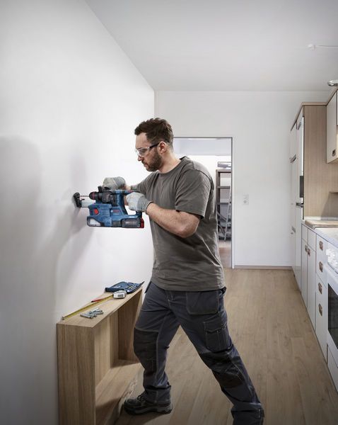 A person wearing safety equipment drills a hole into a wall in a kitchen.