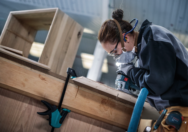 Person wearing safety equipment uses a power tool to shape wood in a workshop.