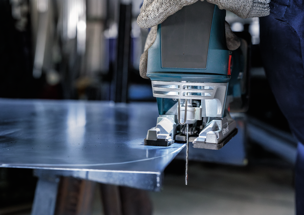 Person wearing safety equipment uses a jigsaw to cut a sheet of metal.