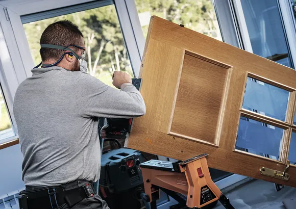 Person wearing safety equipment sanding a wooden door supported on a workbench.