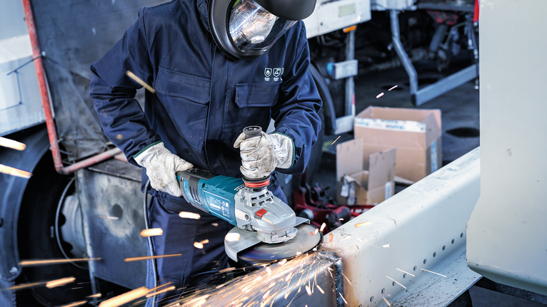 A person wearing safety equipment grinds a metal beam, producing sparks.