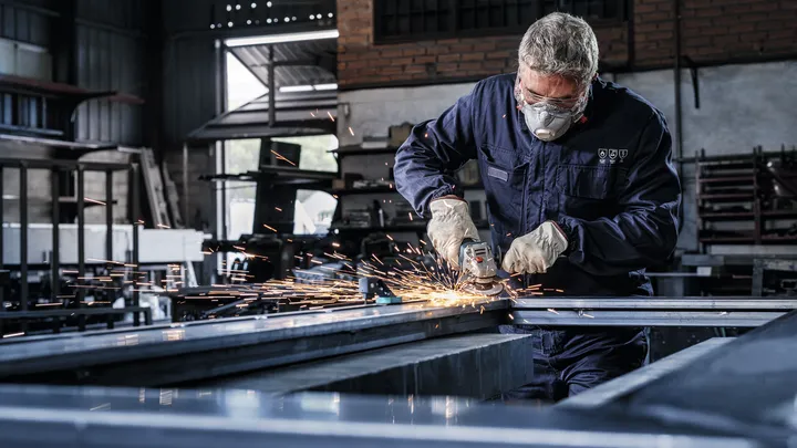 Person wearing safety equipment grinds metal, creating sparks in an industrial workshop.