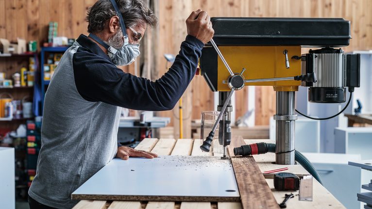 Person wearing safety equipment drills a hole into a board with a stationary machine.