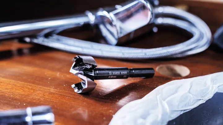 Close-up of a hole saw drill bit beside plumbing parts on a wooden surface.
